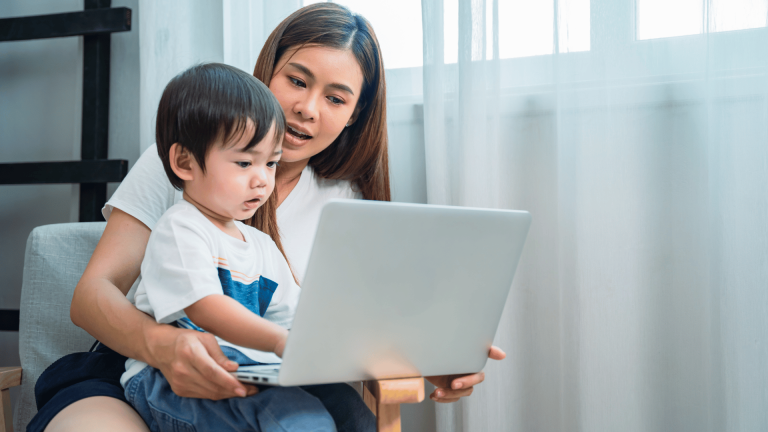 Young mother is sitting in an chair by a window. She holds her child on lap while she has her laptop open